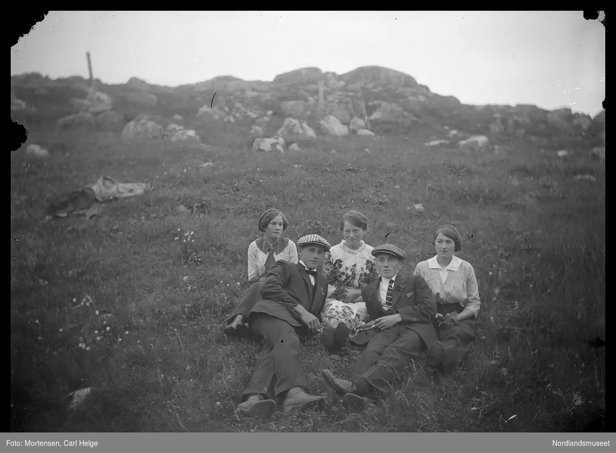 To unge menn i dress og med luer så som tre unge kvinner på en eng, noen bergknauser i bakgrunnen