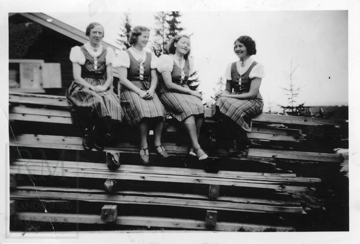 Fire kvinner i blåtøybunad, sittende på en stabel med planker.