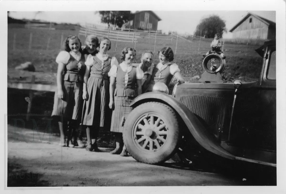 Fire jenter i blåtøybunad og to menn stående foran en bil. Syklubb på tur.