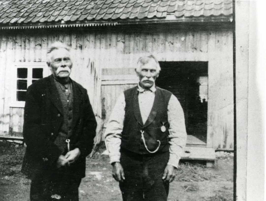 Fotografi av to eldre menn som står foran et uthus eller en låve.