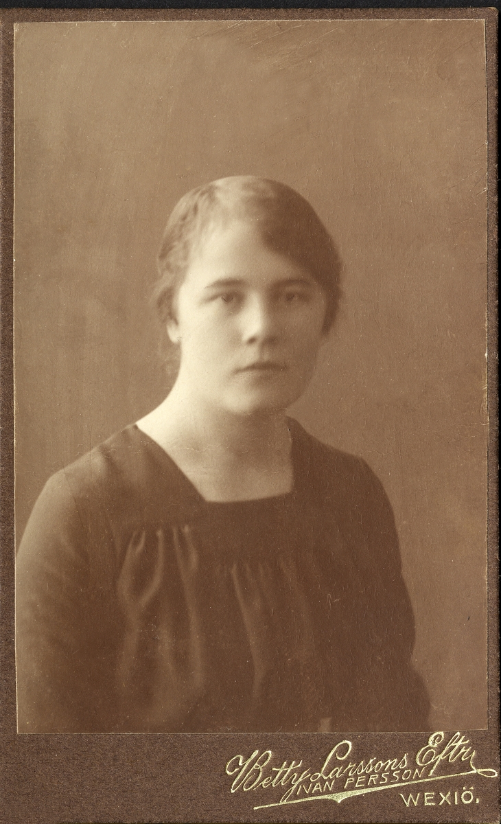 Porträttfoto av en okänd kvinna i mörk enkel blus med smockliv. 
Bröstbild, halvprofil. Ateljéfoto.