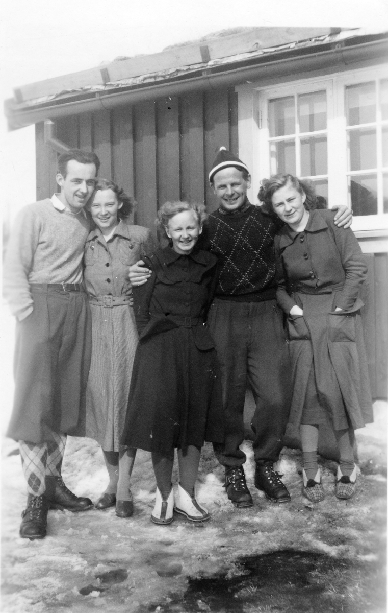 Tre damer og to menn oppstilt foran lite hus