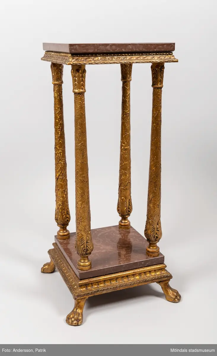 Piedestal från 1920-talet.  Piedestalen är tillverkad av förgyllt trä med rödbruna marmorskivor. Den har fyra stycken rikt utsmyckade ben och står på fyra lejontassar, som också är tillverkade av förgyllt trä.