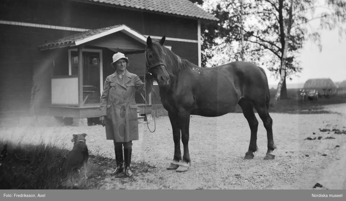 Fogelstad. fröken Anna Rasbo, ladugårdsförman stående  i helfigur framför ett hus med en hund sittande vid sina fötter,  Med vänster hand håller hon i tyglarna till en häst.