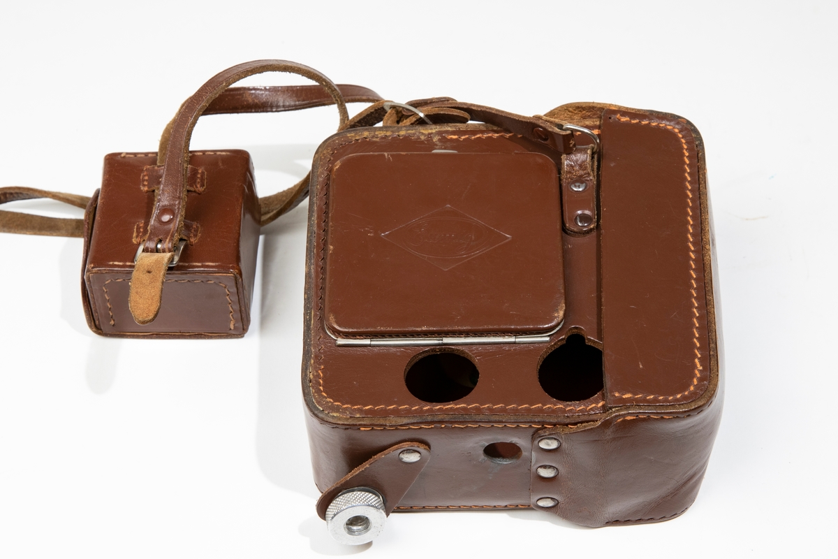 Väska till filmkamera, dubbel-8 Eumig C 3, av brunt läder med öppningsbart lock framtill och lucka på sidan. Väskan har axelrem av läder, på vilken det sitter en mindre fyrkantig väska, innehållande litet linsskydd i en röd tygpåse, och en fjärrutlösare.