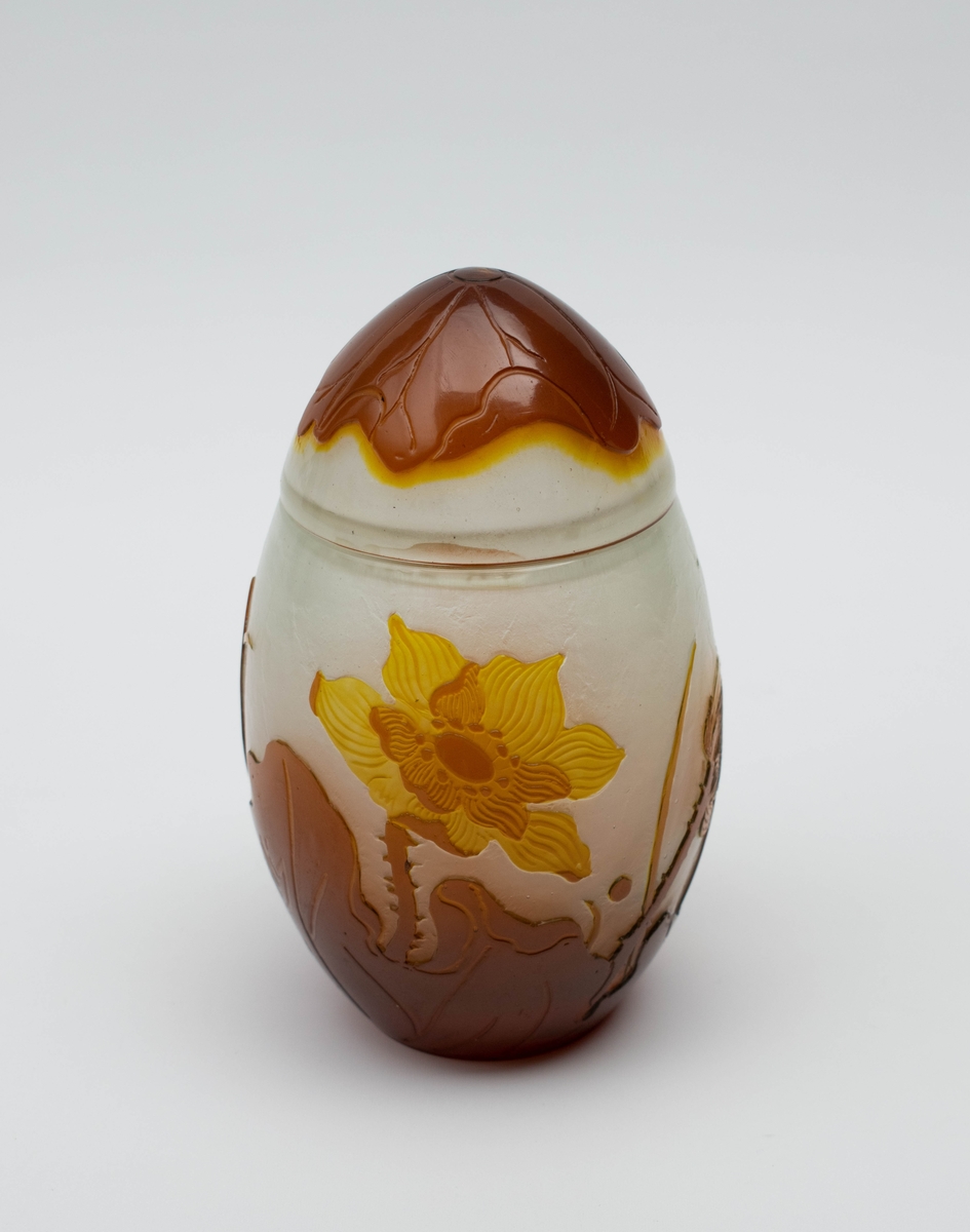 Vase med lokk i melkehvitt opakt overfangsglass. Eggeformet korpus som er dekorert med en gul blomst samt brunlig bladverk i overfangsteknikk. Deler av overflaten er trolig etset.