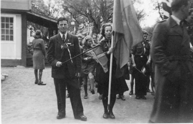 Ett led med scouter: flickor och pojkar. I täten står en ung kvinna med en fana och intill en man i civil klädsel som viftar med en flagga.