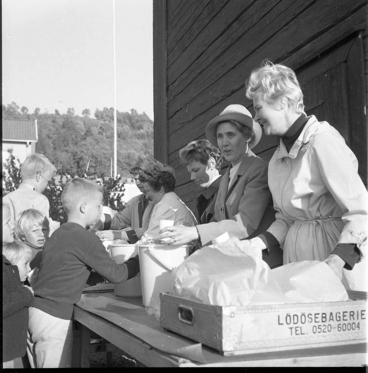 Barn står vid ett bord utmed en husvägg där kvinnor delar ut saft och bullar. På bordet står en trädlåda märkt "Lödösebageriet". Kvinnorna är från höger: Birgit Roman och Birgit Heldebrandt