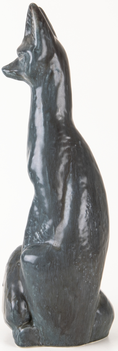 Figurinen Räv av formpressad flintgods, formgiven av konstnär Lillemor Mannerheim. Grågblå glasyr. Sittande räv med svansen lindad runt nedre delen av kroppen.