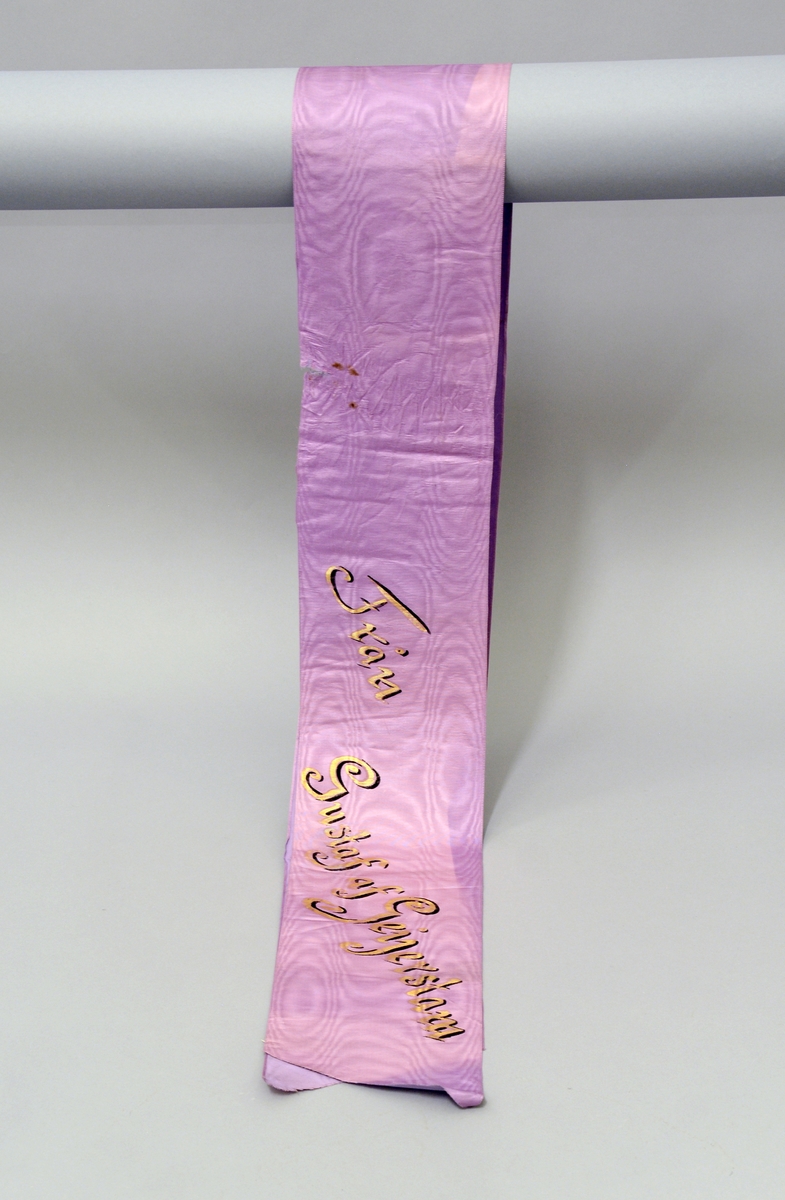 Båndet er lilla (rosa) med gulltekst - håndskrevet. 