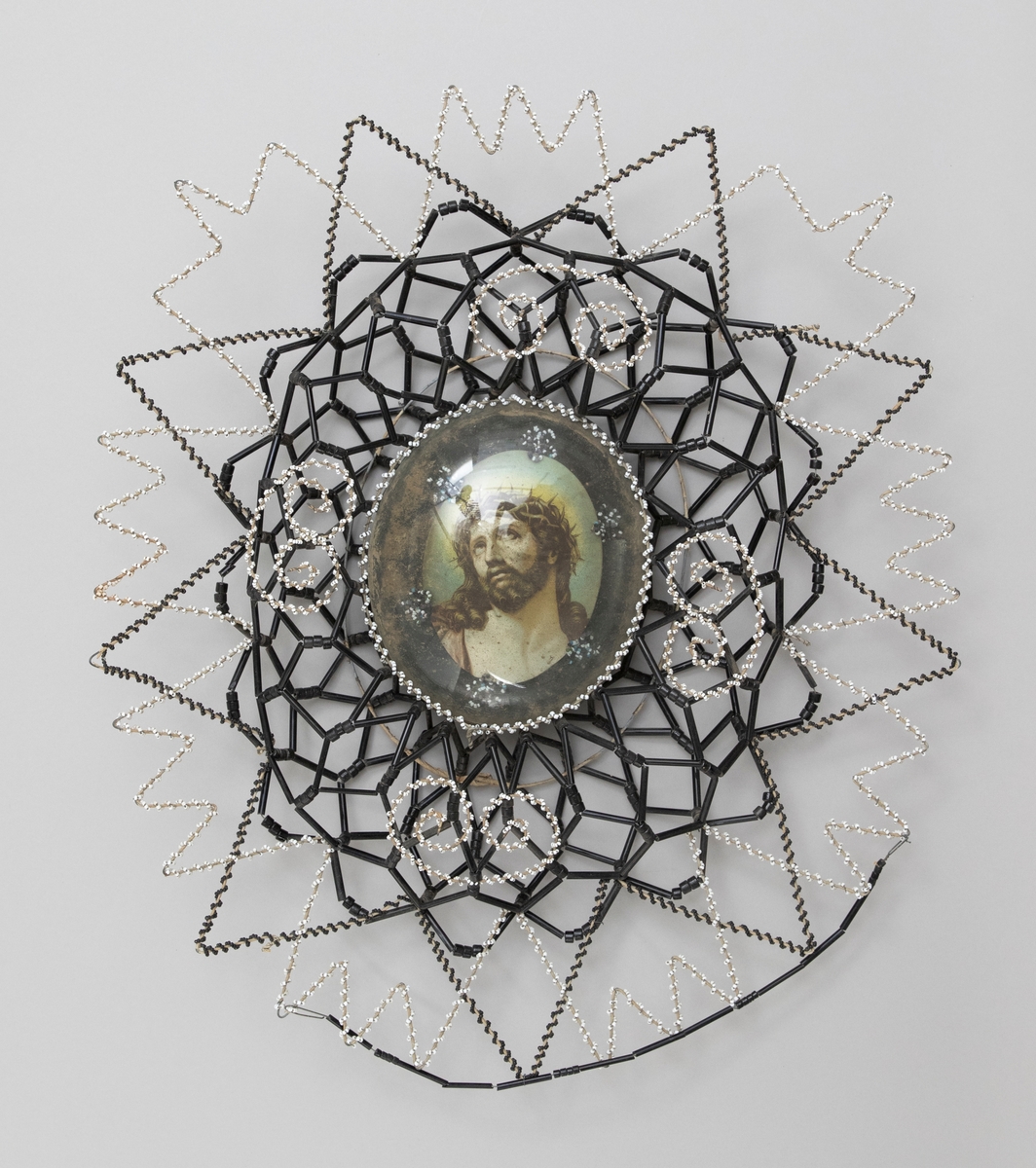 Pärlkrans på stomme av järntråd med dekorativt mönster. I mitten en Kristusbild under ovalt, kupigt glas, däromkring en krans av svarta och vita glaspärlor.