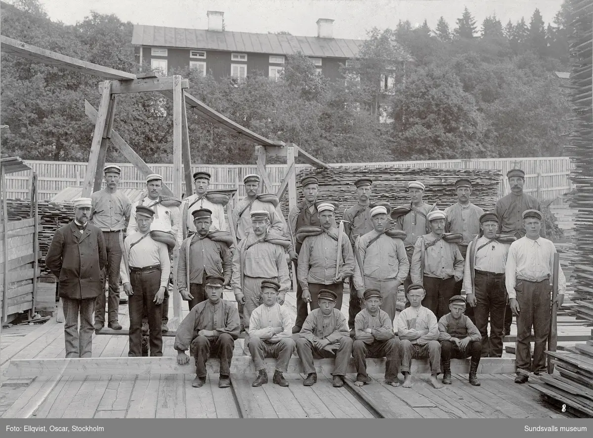 Brädgårdsarbetare, Fagerviks sågverk. I bakgrunden en av arbetarbostäderna. Per Aron Landin  (f. 1867 d. 1946) står i översta raden 4:e man från höger. Landin blev senare handelsföreståndare i Fagervik.