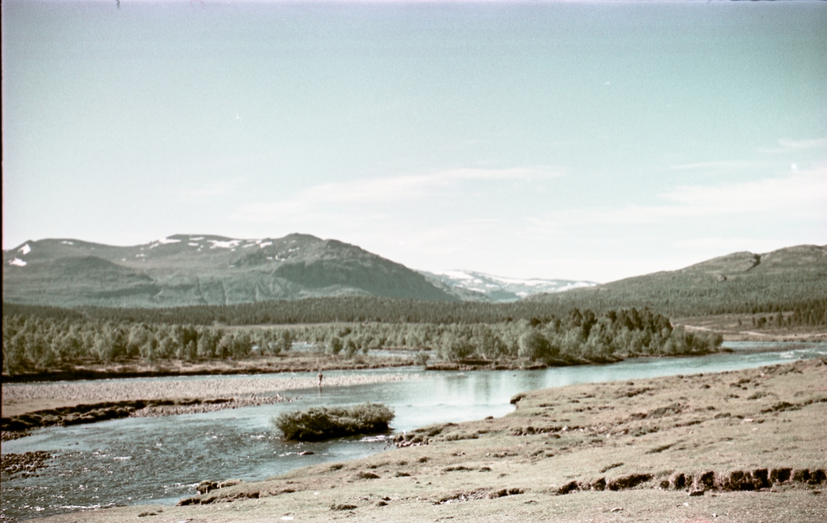 Landskap med elv i forgrunnen, fjell i bakgrunnen. Bildet er trolig tatt et sted mellom Maurvangen og Riddersparanget, og elva er Sjoa.