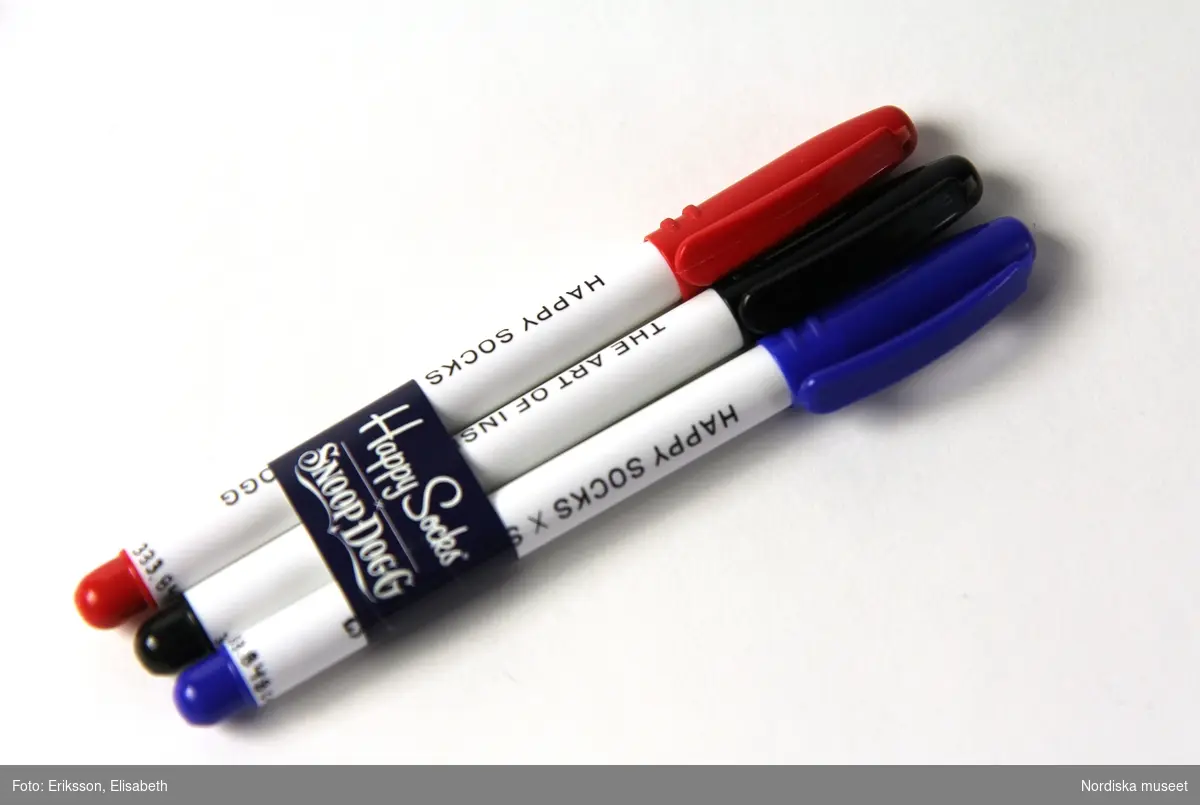 Tre tuschpennor i plast (A-C). Runt pennorna är en bred blank papperslapp (D) fäst med tejp för att hålla ihop pennorna. På lappen står det ”Happy Socks x SNOOP DOGG” i vitt. 
Varje penna är i vit plast med ett löstagbart kork i en annan färg som symboliserar dess tuschfärg; en röd, en svart och en blå. Den rundade änden längst ner på varje penna har också denna färg. På det vita partiet står det ”HAPPY SOCKS X SNOOP DOGG” på ena sidan och ”THE ART OF INSPIRATION” på andra sidan. Varje pennkork har en klämma.
 /Elise Wargentin 2015