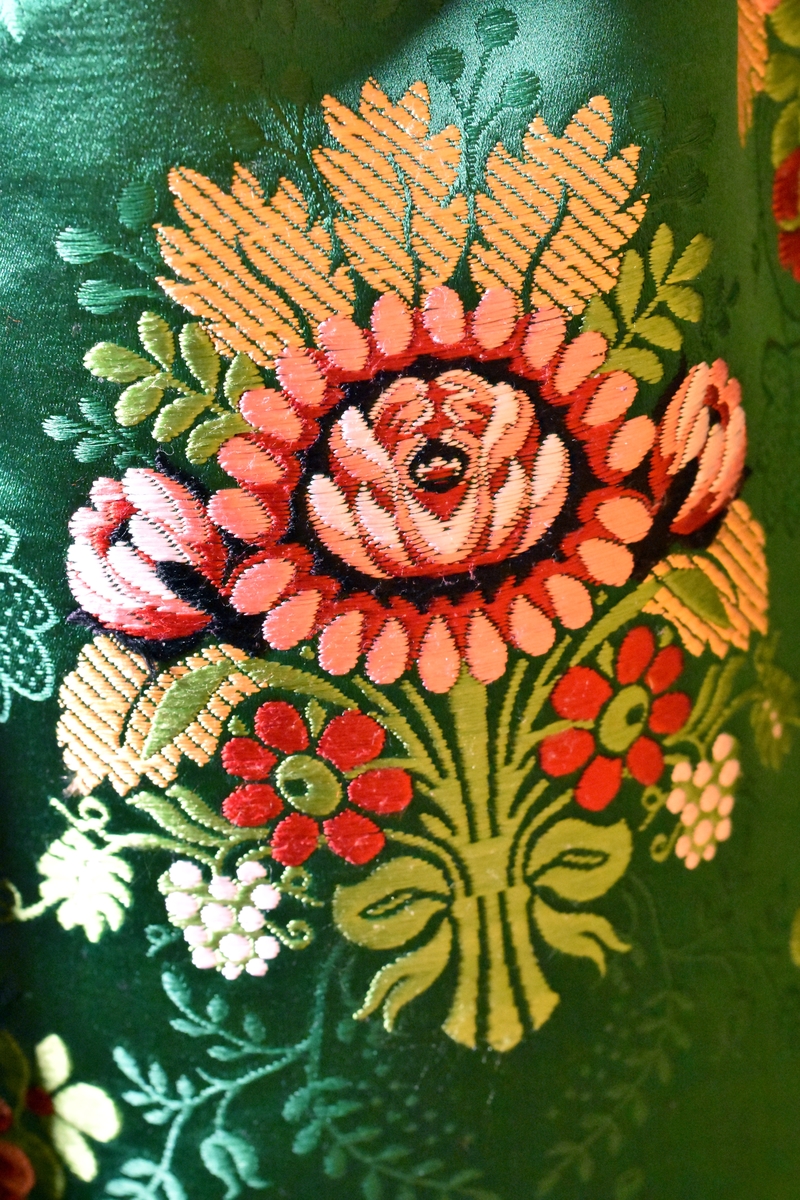 Förkläde i sidenbrokad, grön botten med stora inplockade rosenbuketter i flotterande silke i rött, rosa, ljusgrönt, vitt och brunt. Upptill dragsko med iträtt grönt sidenband. 
Förklädet är skarvat utan mönsterpassning på ena sidan. Kanten är skodd med grönt sidenband från "Mokuba". Skarvarna är sydda på maskin. I övrigt är plagget handsytt.