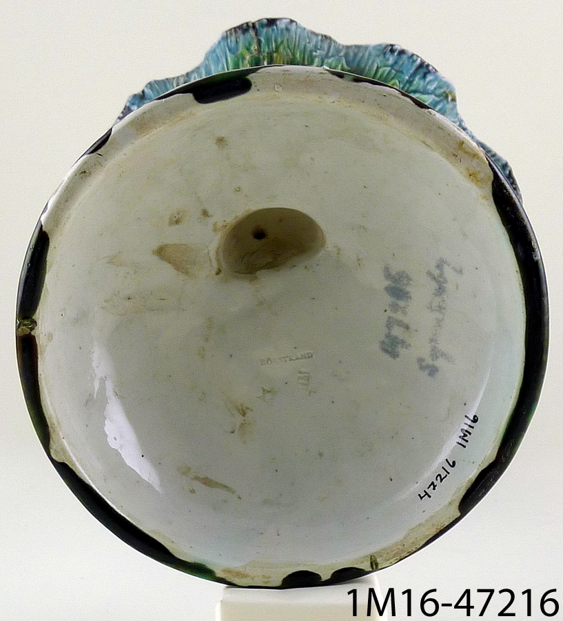 Skål, i form av gosse stående på en kulle, bärande upp en skål i mussleform. Föremålet är handmålat i blått, beige, brunt och grönt.