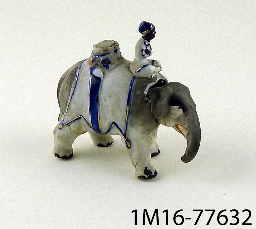 Tvådelad figur i porslin föreställande elefant med elefantförare. Figuren ser ut att ha varit konstruerad så att elefanthuvudet varit rörligt.
