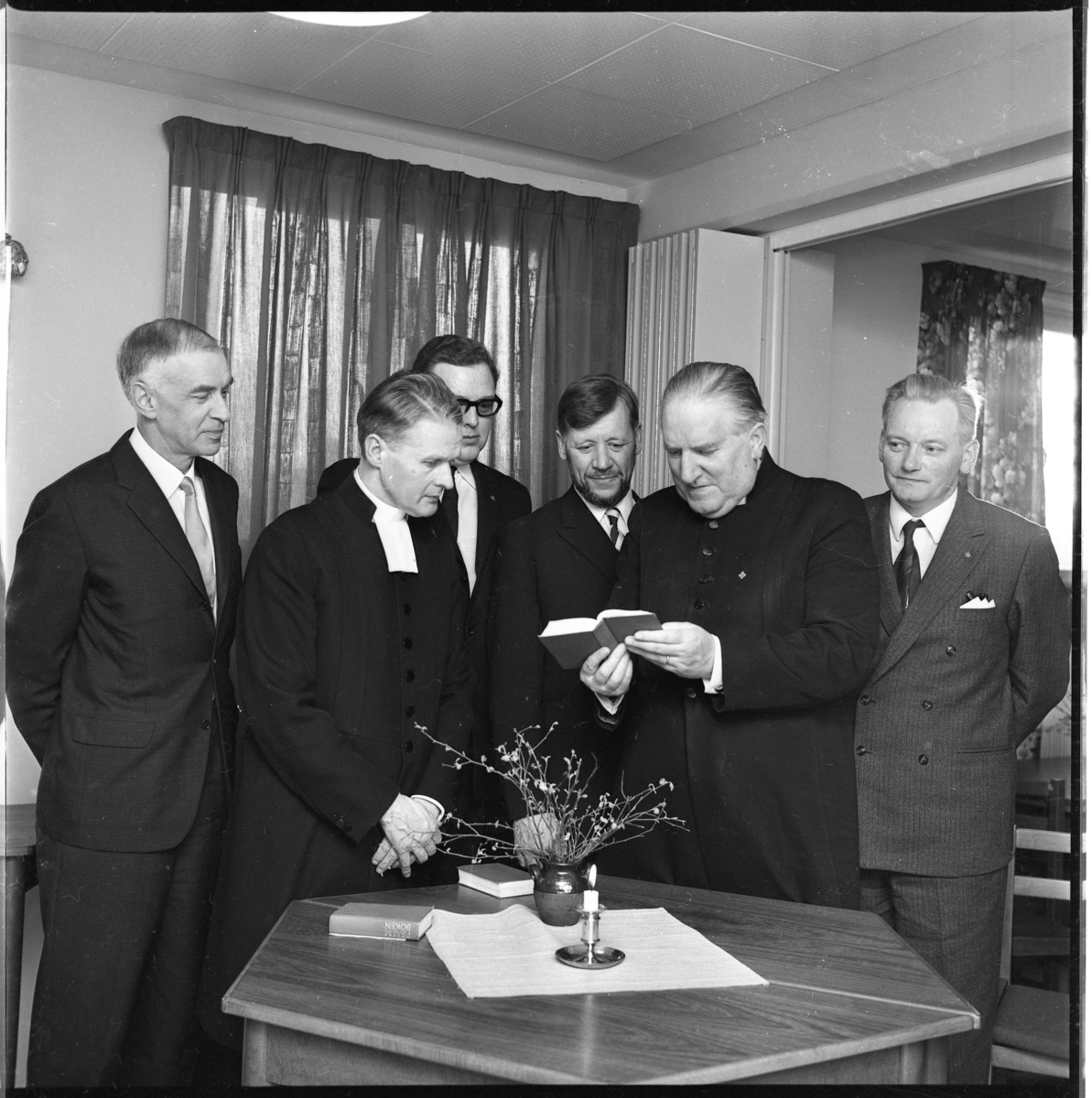 Sex män varav två prästmän samlade runt ett bord med ett tänt ljus. Man tittar mot, troligen en psalmbok som kyrkoherde Wallis Wendel håller i händerna. De är från höger Göran Fong, Wallis Wendel samt Per-Axel Wenner.