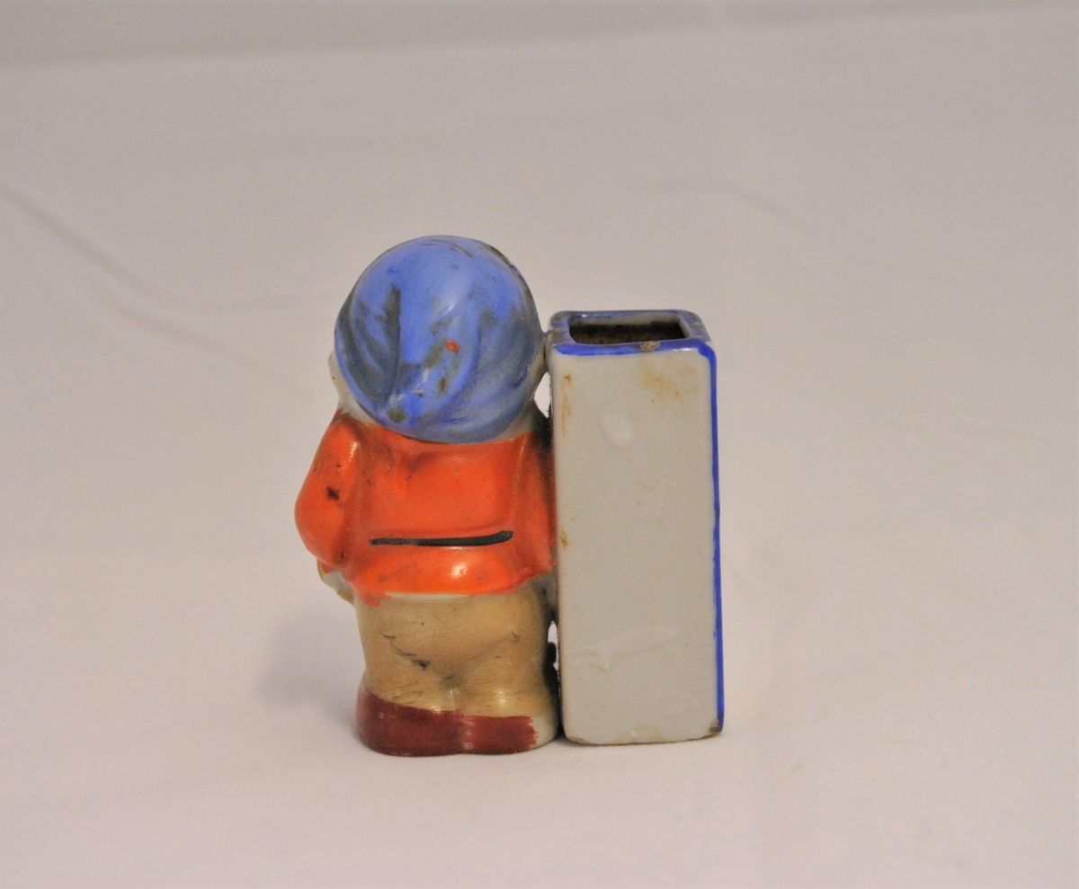 Liten figur i form av en av dvergene fra Snehvit (Blygen?) i porselen. På sin høyre side har han en firkantet beholder, mest sannsynlig til oppbevaring av tannpirkere.