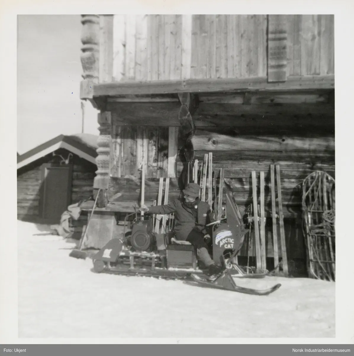 James Coward på snøskuter foran loft. Ski står stablet langs loftsvegg, Ellefstjønnburet