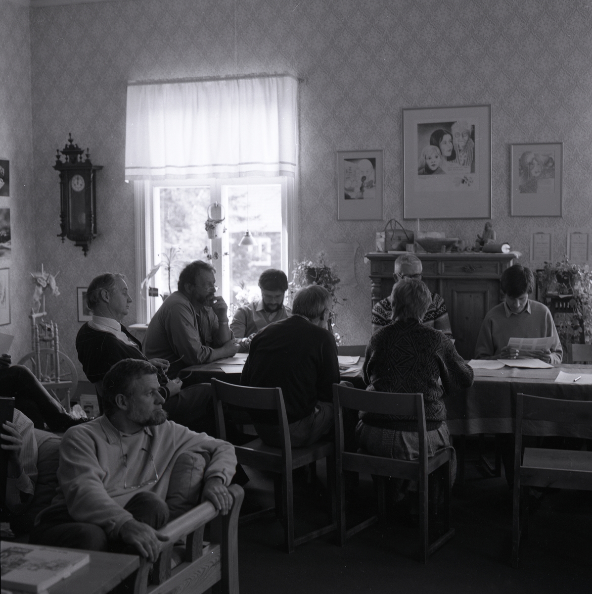 Inne i huset sitter det män och en kvinna på stolar runt bordet, dom läser i papper. I fönstret är det en gardin, blommor och en lampa. På väggen hänger en klocka och tavlor, på golvet står en spinnrock och en gammal skänk. 25-26 april 1987.