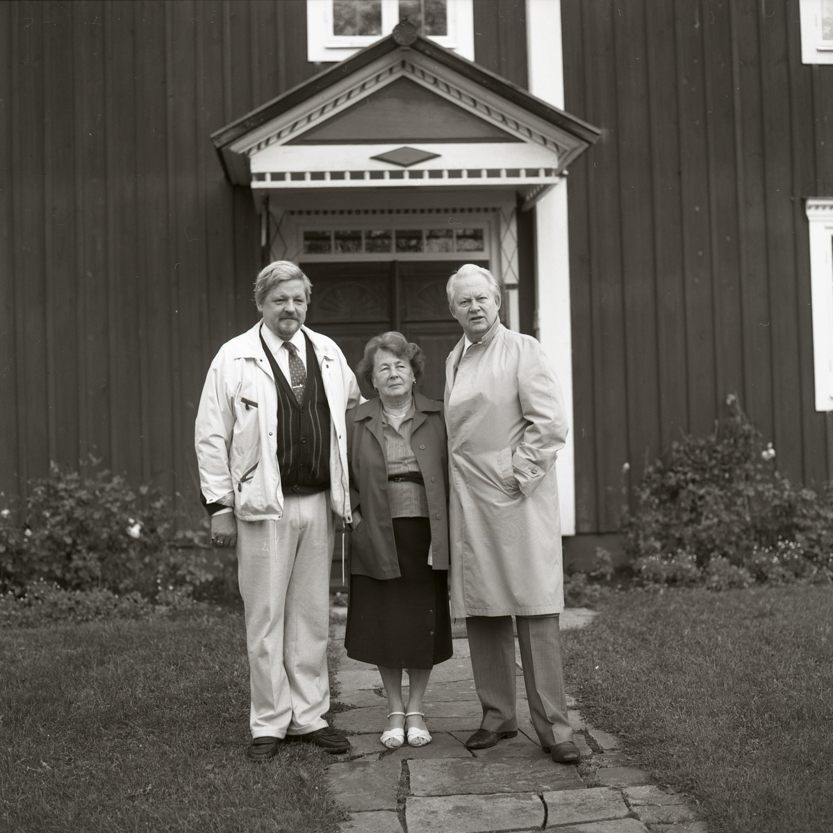 På stenplattorna som går till huset står två män och Adéle står i mitten. Bakom är ett hus med fönster, dörr och ett tak ovanför dörren med snickarglädje. På marken är det gräs och blommor växer mot husväggen. 30 september 1990.
