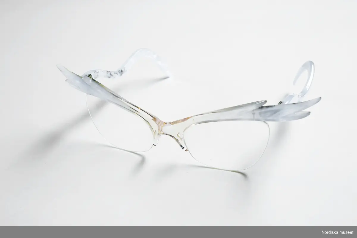 Ett par glasögon med pärblå "vingad" kant, troligen av plexiglas. Nylontråd fungerar som en kant under glasen. Trådarna är fästa vid näskuddarna samt på frontens baksida, resten av trådarna (ungefär 7 cm) hänger fritt på baksidan. Oslipade glas med sadelbrygga. Vågformade skalmar i samma pärlblå färg. 
/Petrine Knight, 2015-07-01