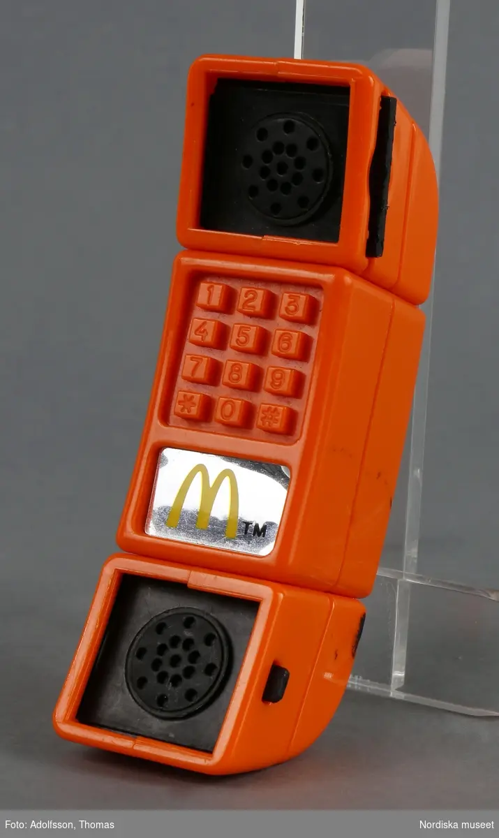Optisk leksak i form av en orange mobiltelefon med 12 fasta knappar med siffrorna 0-9 samt * och #. De svarta plattorna för lur och högtalare kan skjutas ut åt sidorna och om man kikar in i hålen ser man en spegelbild av sig själv och får en känsla av att mobiltelefonen förvandlas till en kikare.