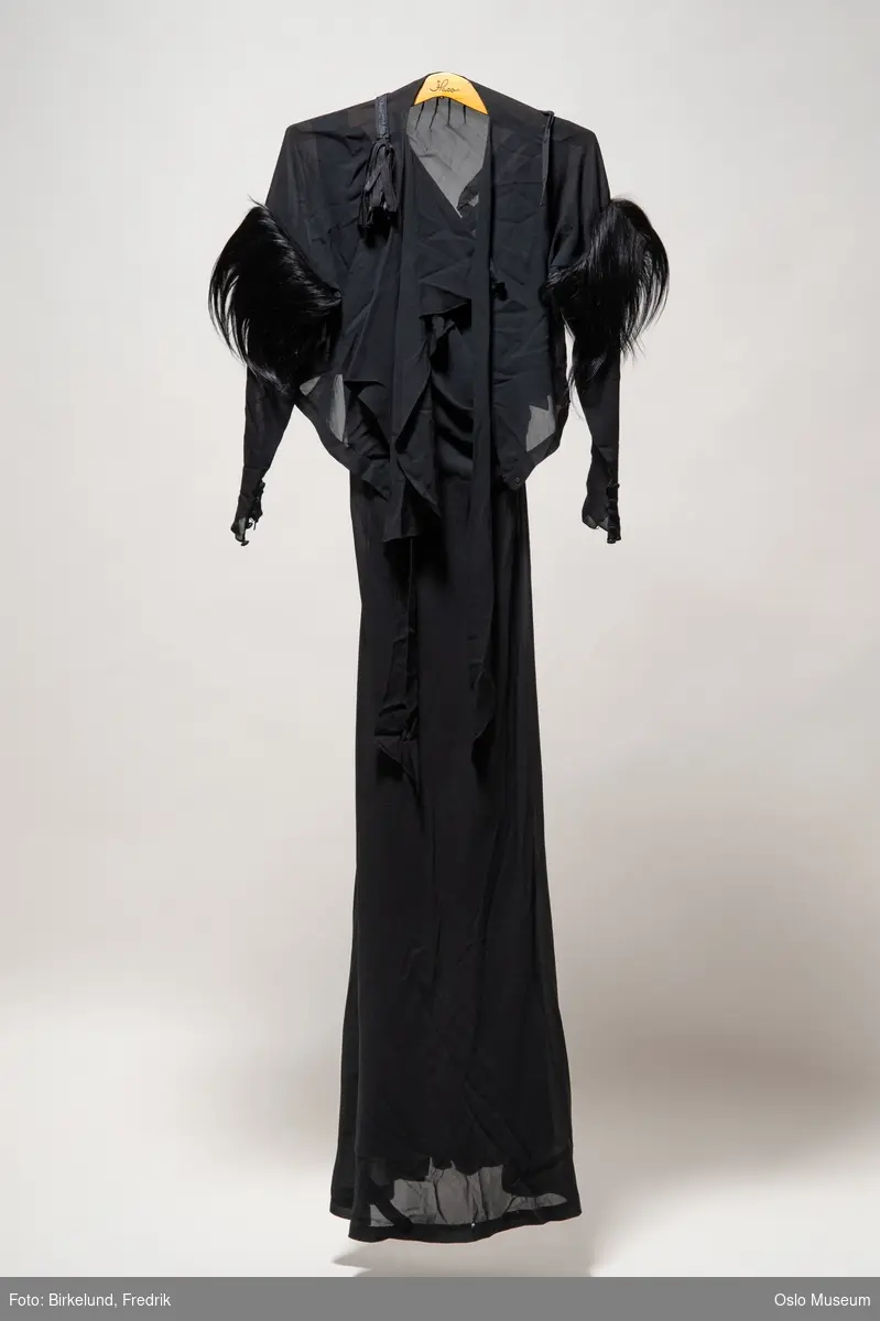A: lang sort ermeløs kjole, 
B: bolero med pelsbesetning på ermer. Tekstiltrukne knapper langs håndledd. Knytes med bånd i halsen. 
C: belte med dusk