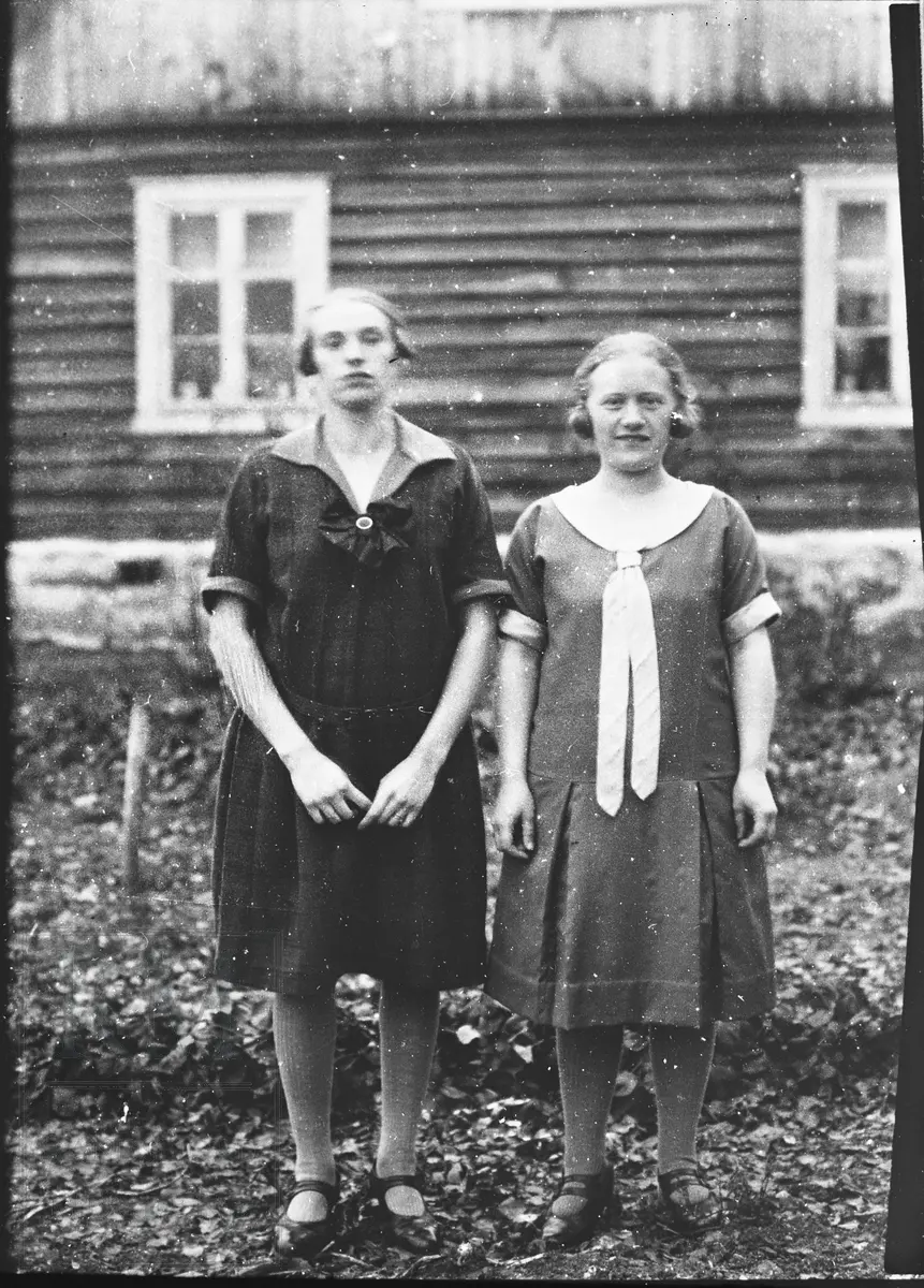 To pyntede jenter står ute foran ei tømmerstue.
