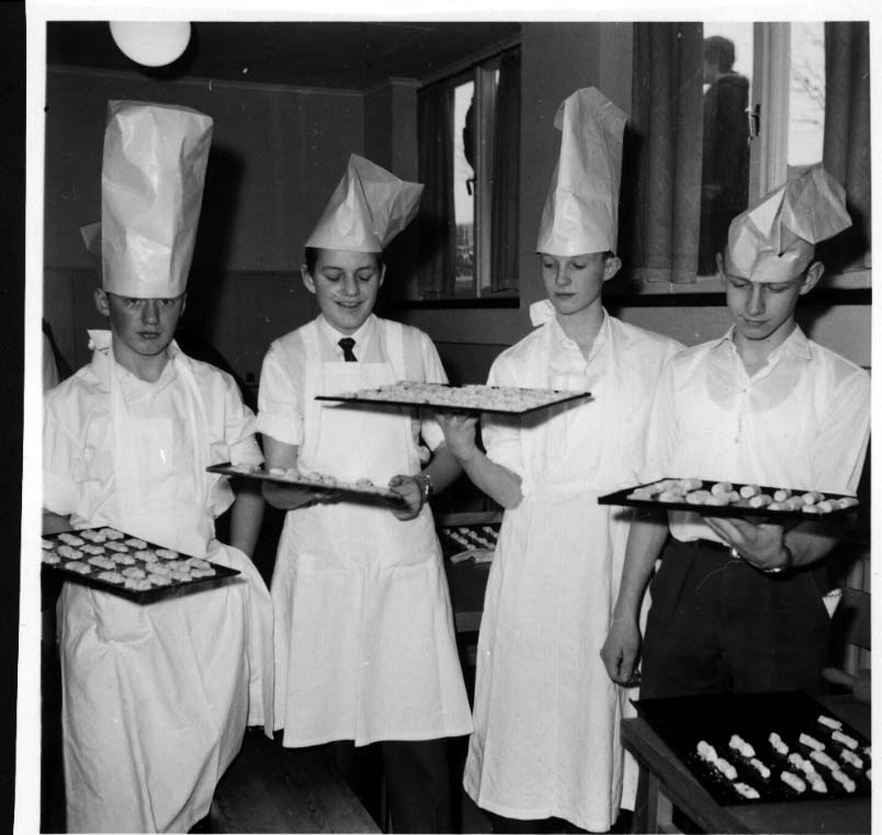 Fyra pojkar varav tre med förkläde håller fram bakplåtar med kakor. Alla pojkarna har en upp och nedvänd papperspåse på huvudet, likt en kockmössa.