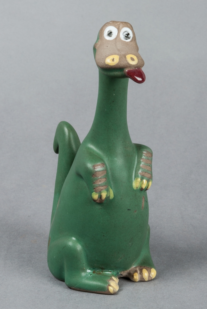 Figurin Dino. Gefle Porslinsfabrik 1957, formgivare Dorothy Clough. Sittande dinousarie med utsträckt tunga. Figuren ingick i den amerikanska serien The Flintstones.
