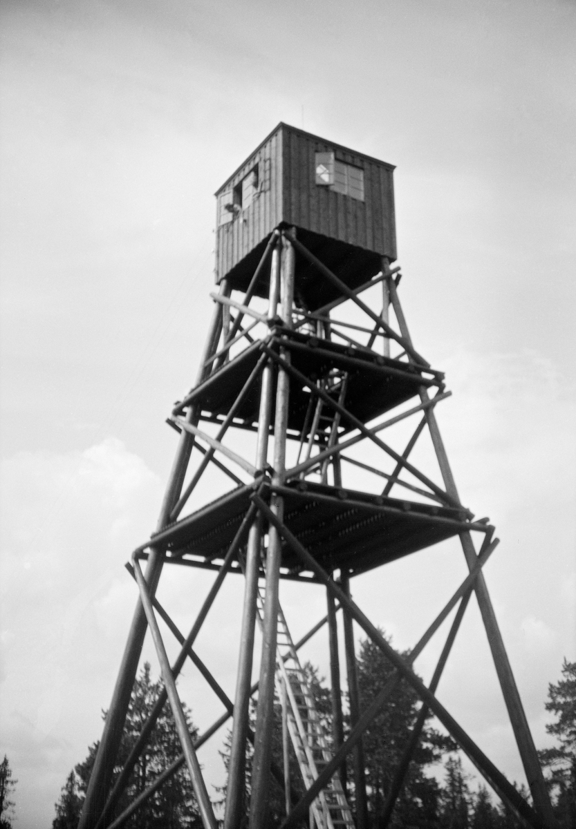 Tårnet på skogbrannvaktstasjonen Haukåsen øst for Lutvann i Østre Aker (Oslo).  Det første anlegget her ble reist i 1911 av Aker kommuneskoger.  Et nytt anlegg finansiert av forsikringsselskapet Skogbrand, men fortsatt eid av Aker kommuneskoger, ble reist i 1934.  Det er antakelig dette andre anlegget som er fotografert på dette bildet.  Tårnet var en stolpekonstruksjon av den vanlige typen med noenlunde kvadratisk grunnplan og bratt, pyramidal form.  Bærekonstruksjonen består av hjørnestolper - montert i par nederst, enkle lengre oppe - avstivet med stolpekryss mot sidene i tre nivåer.  Mellom hvert nivå er det lagt golv eller plattformer på horisontale stolpelunner.  På toppen av tårnet er det ei utkikkshytte med bordvegger og vinder i alle himmelretninger.  