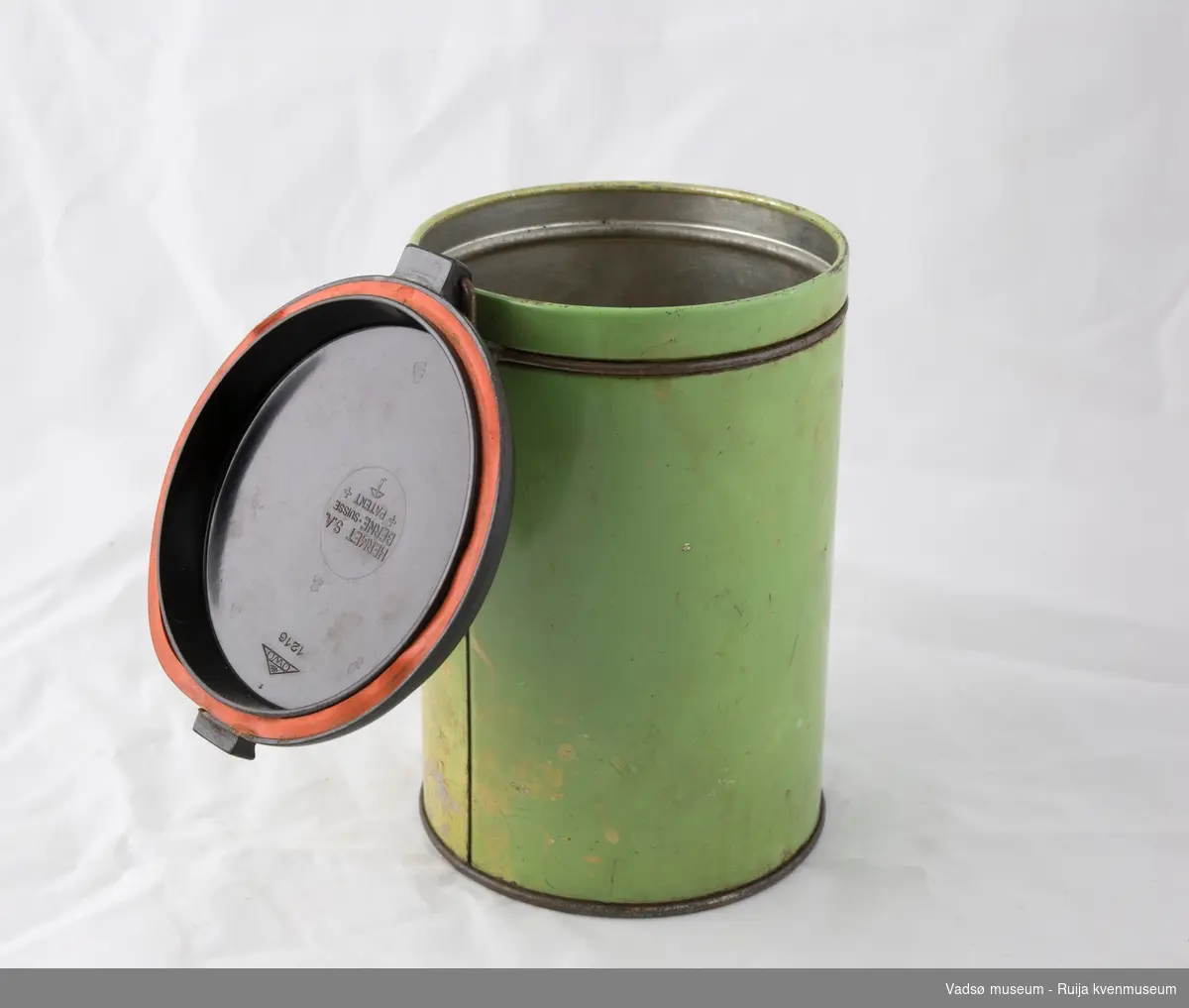 Sylinderformet kaffeboks i metall med hengslet plastlokk. Enkel låsemekanisme i metall i front. Lokket er i brun plast og utstyrt med en gummipakning. Boksen er helfarget grønn utvendig, med påskriften "Kaffe"i svart i front.