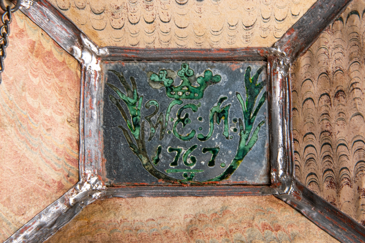 Syskrin. Överst grön nåldyna. Formen är pulpetartad, sidorna bestå av målade glasskivor med blylister emellan. Målningen är grön med blomornament i svart och silver. Gult papper under. Locket rör sig på två gångjärn och hålles innuti av en tunn kedja. I locket under nåldynan finnes en liten spegel med målning i grönt "W.E.M. 1767", med kunglig krona över samt växtranka omkring nertill. Skrinet står på fyra små kulfötter av bly. På ena fältet av locket ser man glasmästareämbetets sigill. Gävle, Gästrikland.