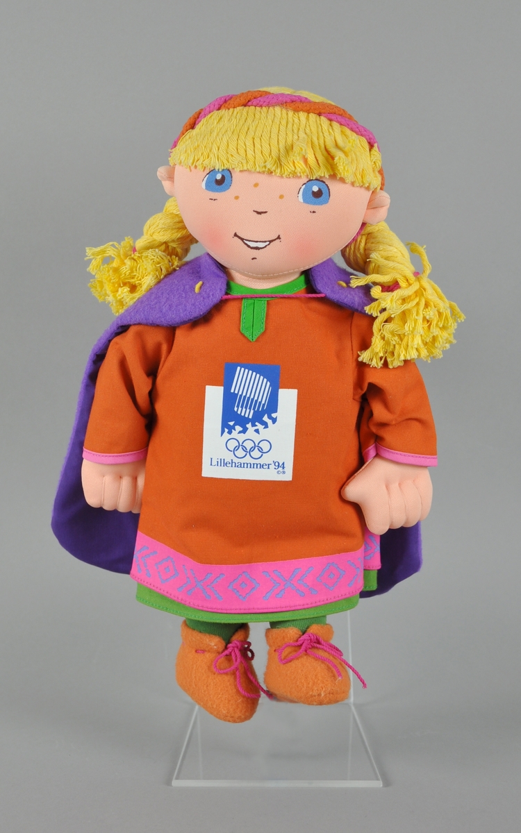 Tekstildukke som forestiller maksoten Kristin. Dukket har gult hår, hårbånd, lilla cape, oransje kjole med logo for Lillehammer '94 og grønne strømper.