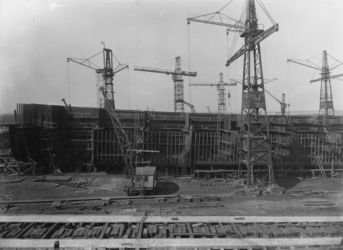 Motivet viser MT SPINANGER (bygget i 1927) under bygging ved Schiechau i Gdansk. 6 store havnekraner rundt skroget som ligger i tørrdokk.