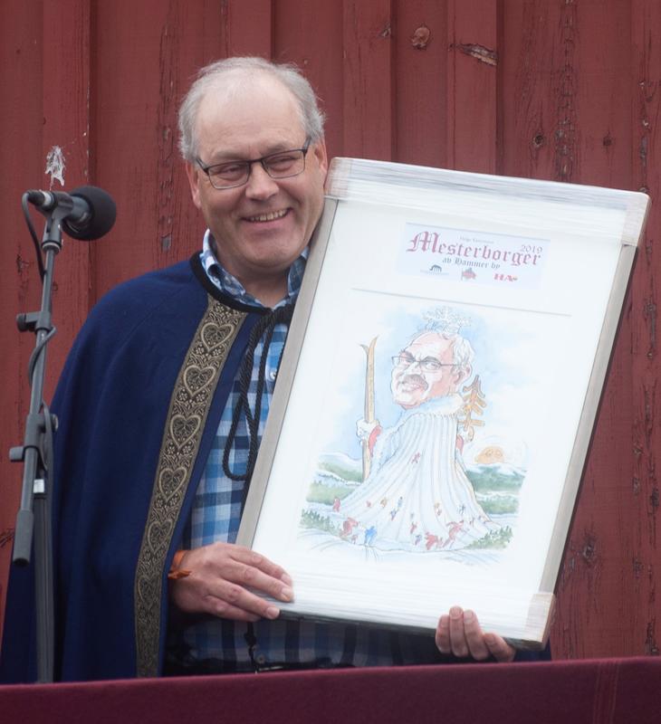 Helge Kartomten årets mesterborger 2019. Mann i blå ullkappe med gullbånd. Holder karikaturtegning av seg selv.