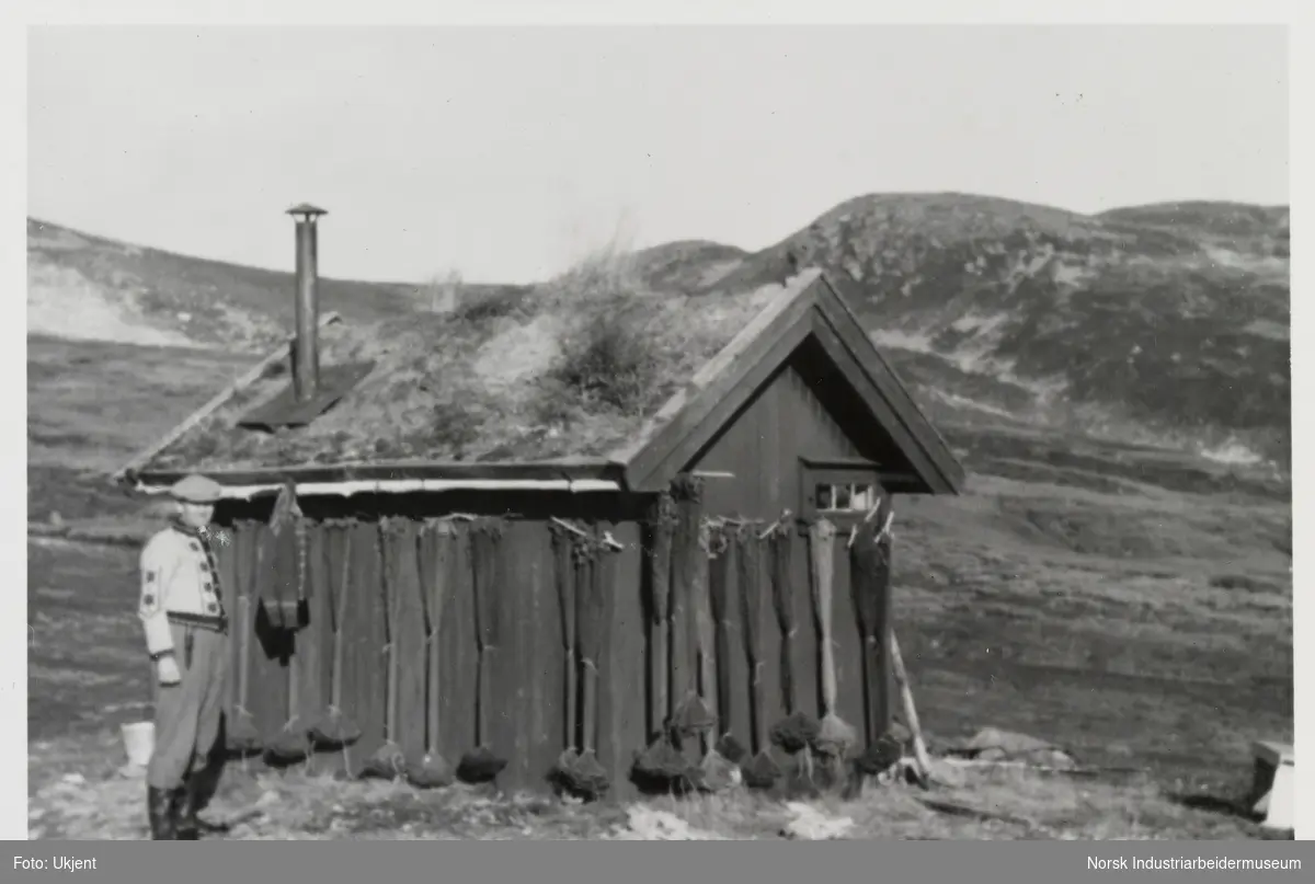 Mann står ved liten hytte i fjellet. Fiskegarn er hengt langs hyttevegg