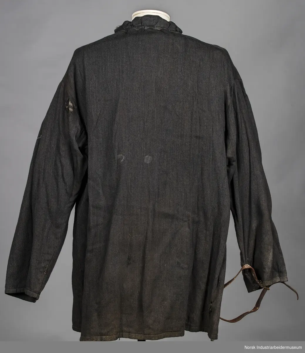 Arbeidsskjorte fremstillet av sort vevet stoff som er lukket med snøre i halsen. Der er festet en lærrem med spenne på det høyre erme.