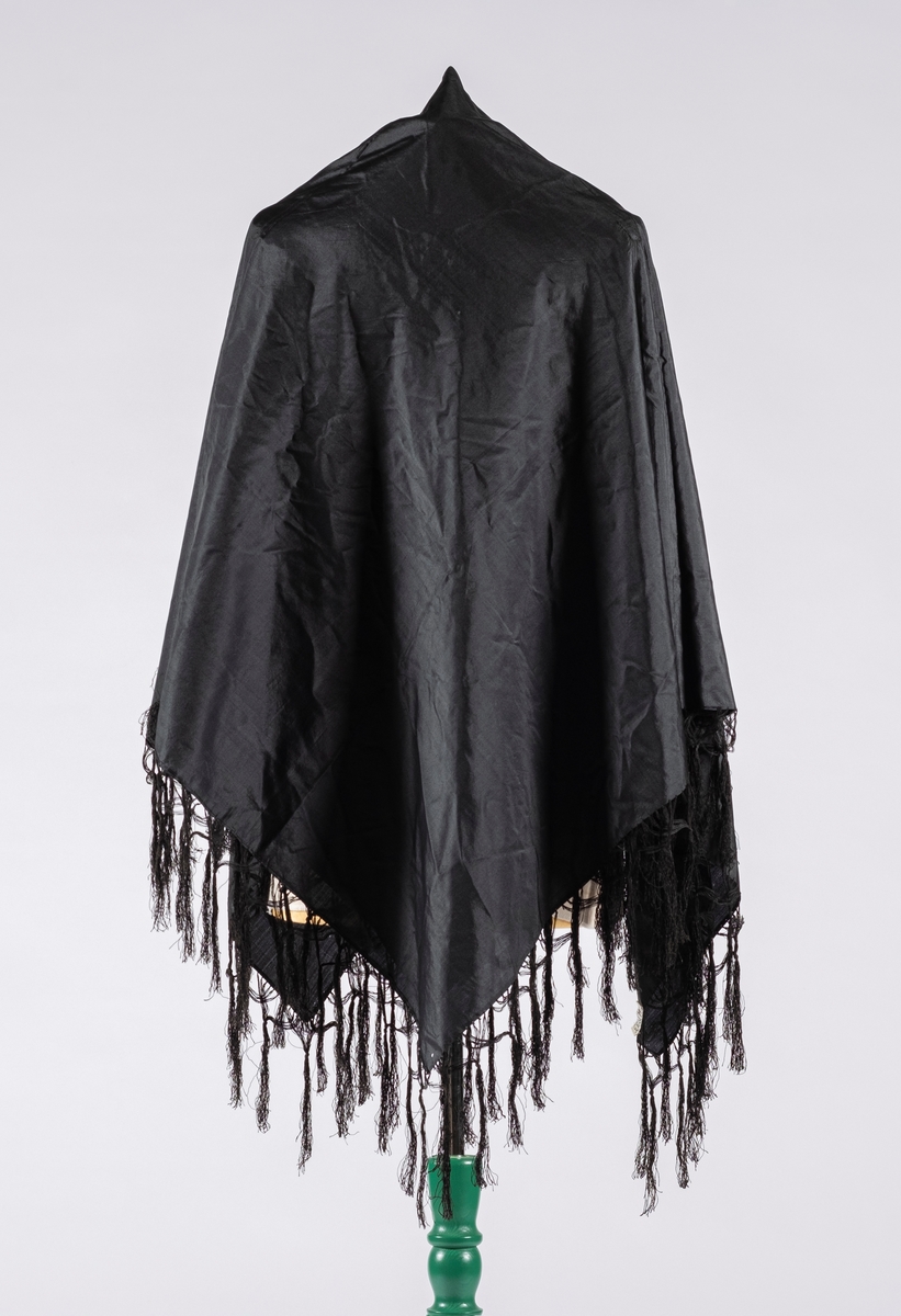 Firkantet svart sjal i taft med knyttet frynsekant. Klippekant i to av endene som er sydd inn.