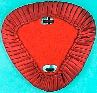 Trekantig mössplåt med rundade hörn, klädd med rött tyg och kantat med veckat sidenband. Hål för fastsättning av mössmärke.