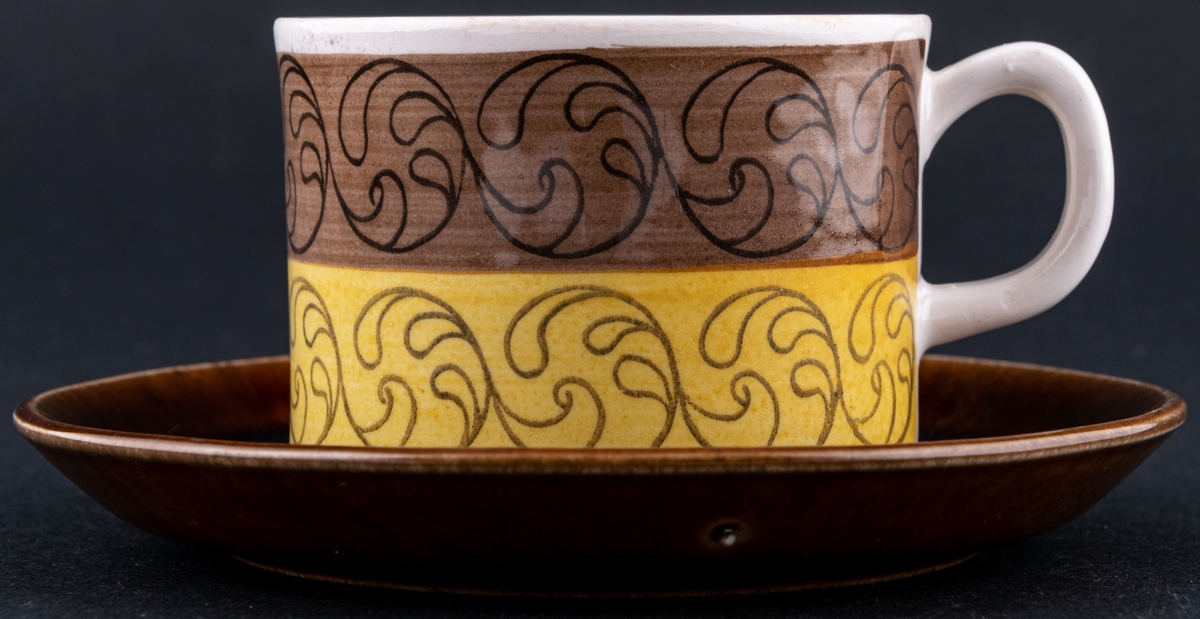 Kaffekopp med fat, modell EH i dekoren Ek. Dekor av Barbro Löfgren-Örtendahl. Runt koppen ett brunt och ett gult band med centrumtryck, fatet helt i brunt.