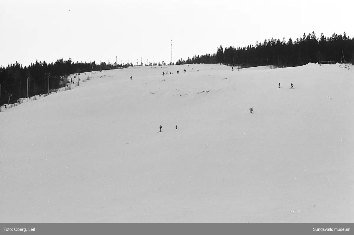 Skönviksbacken med skidåkande ungdomar samt köande liftåkare. I bakgrunden ses Skönviks brukssamhälle.