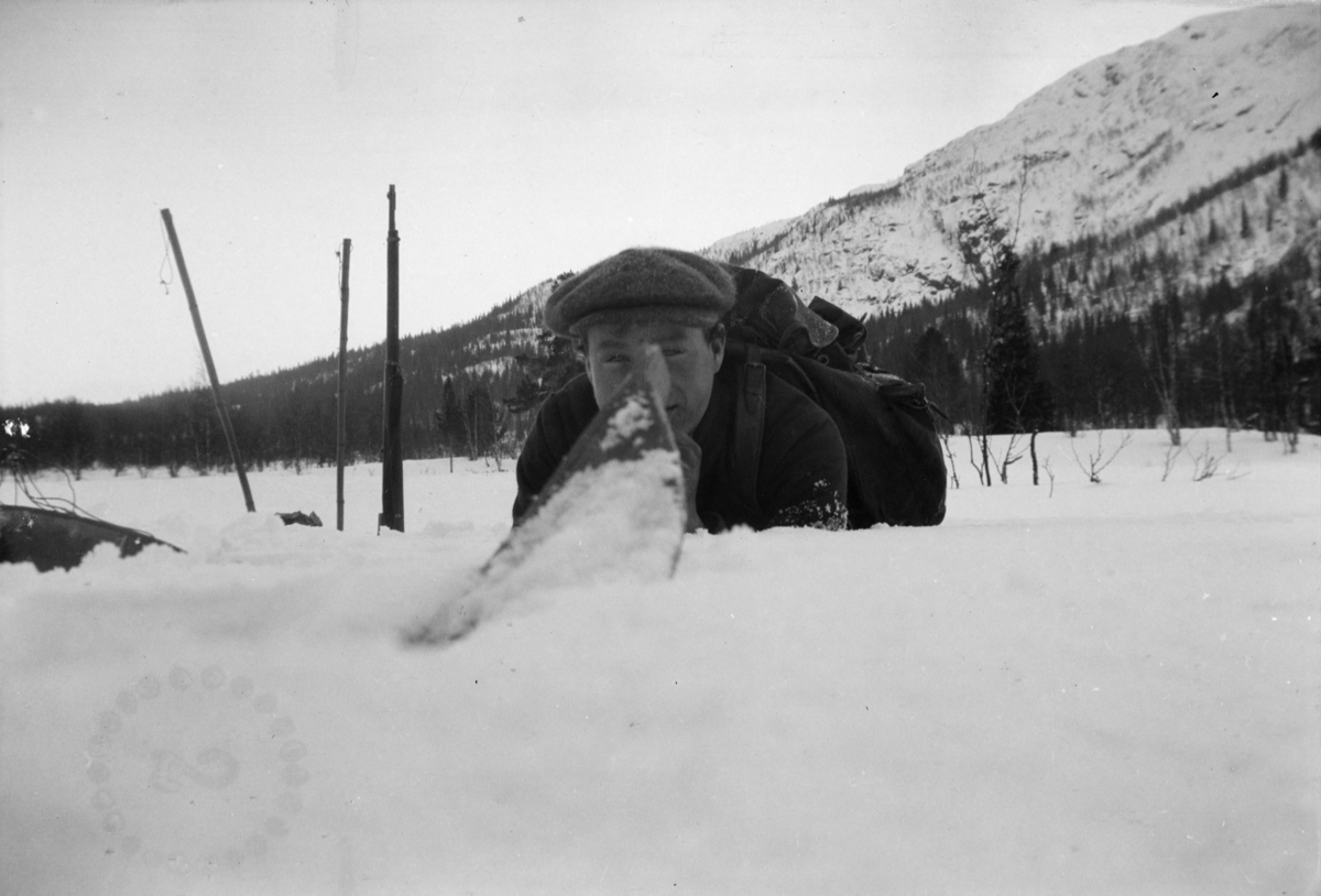 Karl Sannheim ligger i snøen med gevær i bakgrunnen. Et sirkulært merke med tallet 2 inni nederst  til venstre.