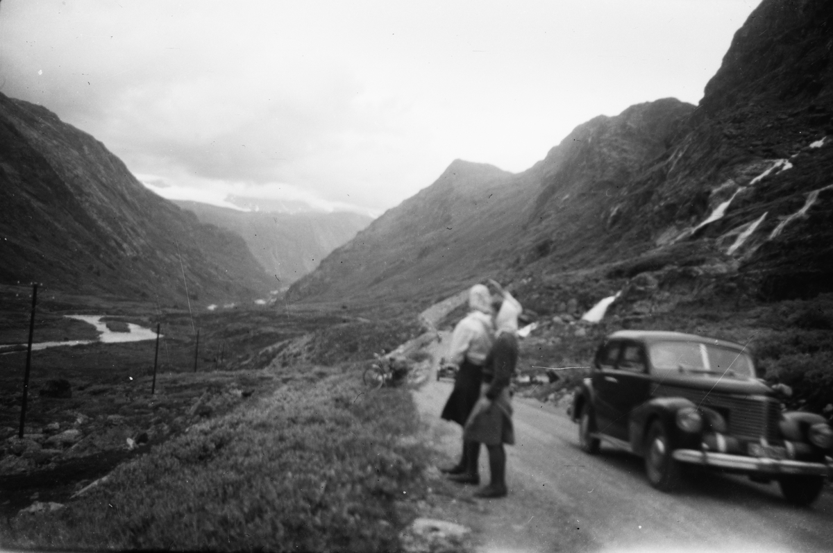 Motiv av to kvinner som står på en vei i en dal og ser på utsikten. På veien er det to biler.