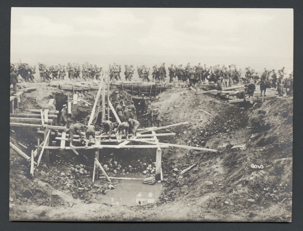 Bilden visar ett stårt hål i marken där en grupp soldater håller på att bygga en träkonstruktion. I bakgrunden syns ett tåg soldater som marscherar förbi.

Originaltext:"Tyska pionierer bygga en nödbro över en mintratt i stridsområdet mellan St. Quentin—Ham."