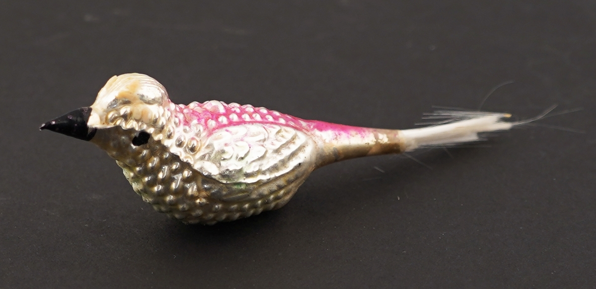 Sølvfarga fugl av glas med rosamåla rygg og blå bringe (slitt) til å sette på juletreet. Stjert av syntetisk materiale. Festeklype manglar