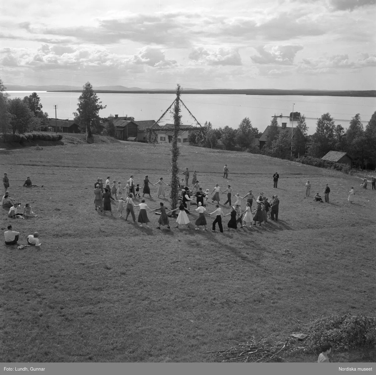 Motiv: (ingen anteckning) (Lerdalshöjden) ;
Tre män spelar fiol vid en midsommarstång, vuxna och barn dansar kring en midsommarstång, en pojke och en kvinna står på en balkong, en flicka - en man och en kvinna sitter vid ett trädgårdsbord under ett parasoll och äter, tomma flaskor av "Loranga" står på ett trädgårdsbord med en midsommarstång i bakgrunden.