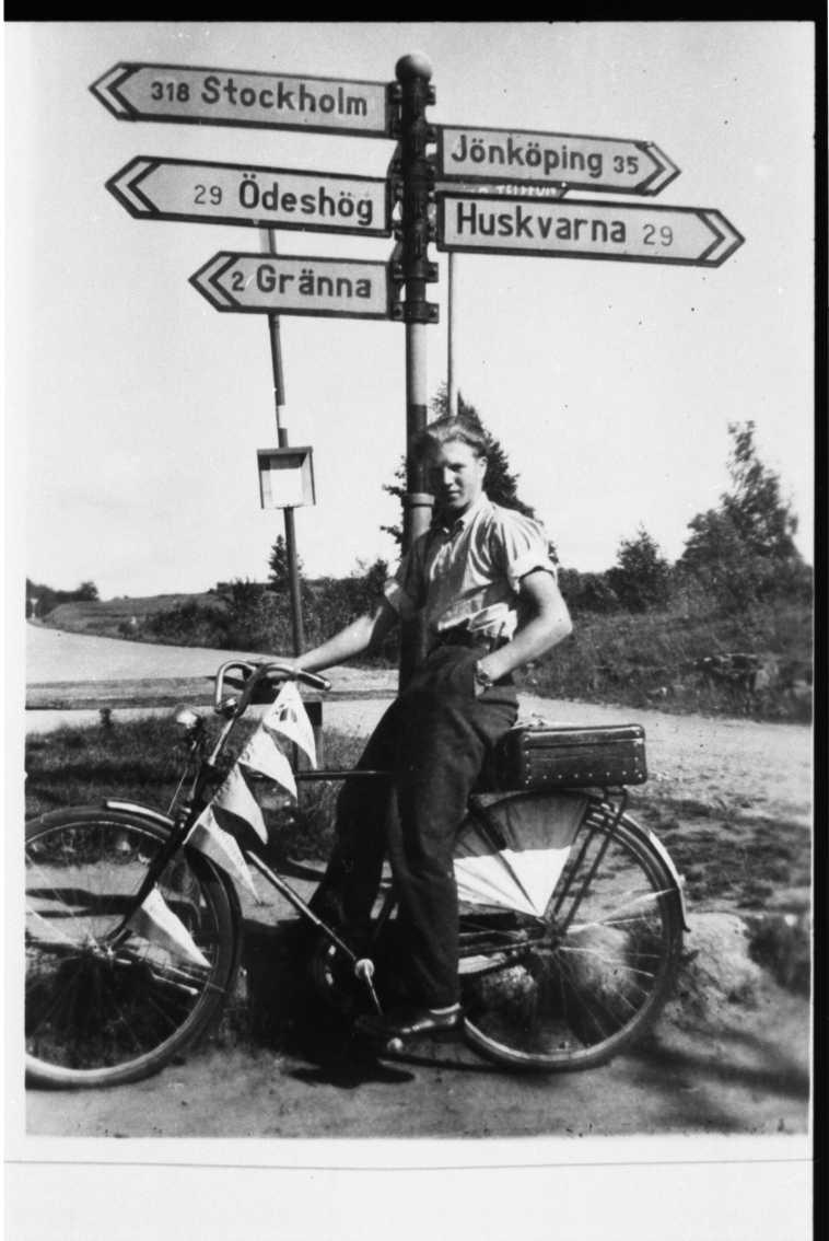 Ynglingen Uno Fransson sitter på en cykel vid trafikplats Galgen söder om Gränna. På pakethållaren har en resväska och från styret till hjulmutter en lina med olika vimplar. Han står vid en vägskylt med hänvisningar till Stockholm, Ödeshög och Gränna mot vänster i bild samt mot Jönköping och Huskvarna till höger i bild.